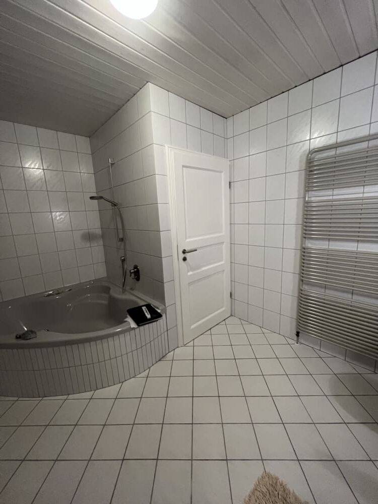 Weiß gefliestes Badezimmer im Deckenlicht mit Eckbadewanne vor der Badsanierung